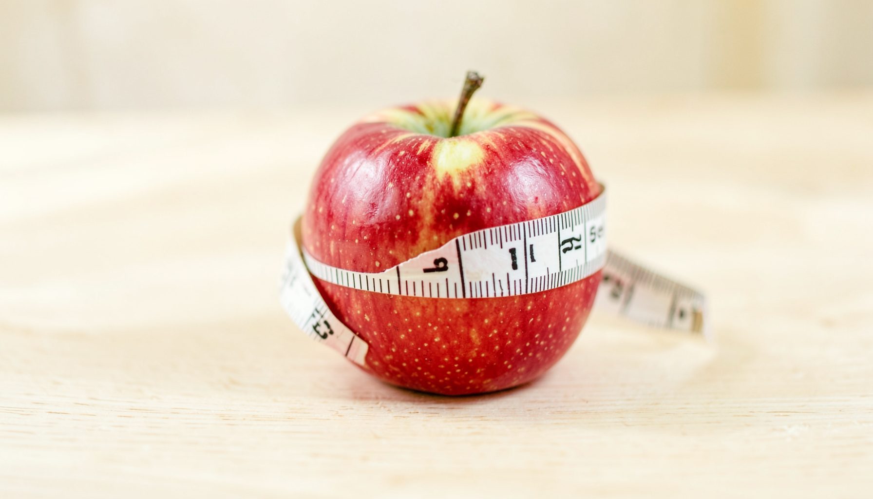 A close-up of a measuring tape wrapped around a fresh apple on a light wooden table, soft daylight, pale cream and red tones.