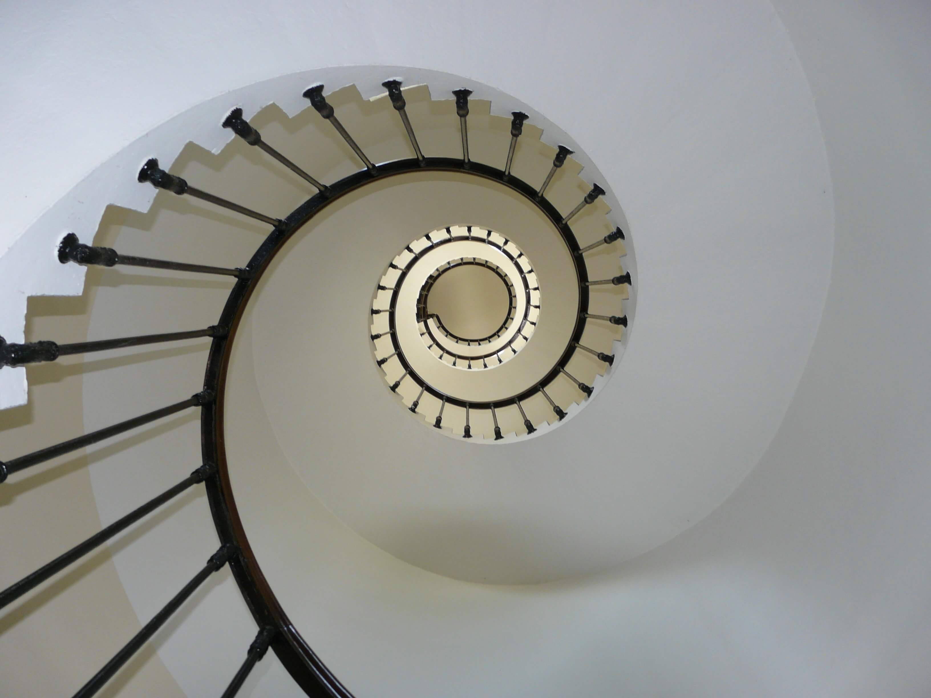 Overhead view of a spiral staircase with dark metal railings, viewed from above showing repeating curves, cream and white tones