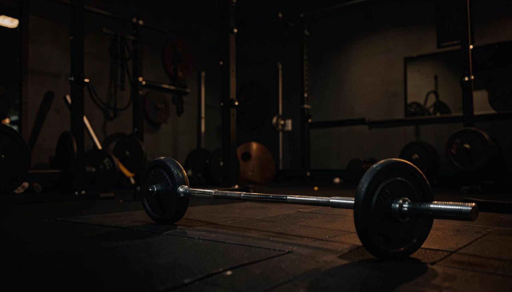 Dark gym interior with weight plates and a barbell on the floor, moody low-key lighting with deep shadows and warm amber accents