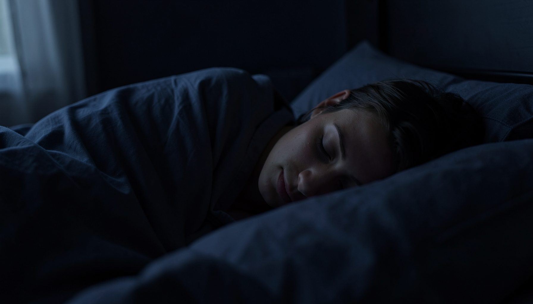 A person sleeping peacefully in a dark bedroom, deep navy and soft grey tones